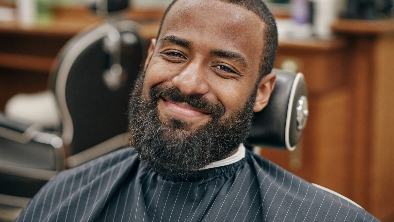 Man in his 30s with a full, well-groomed beard smiling in a barbershop chair