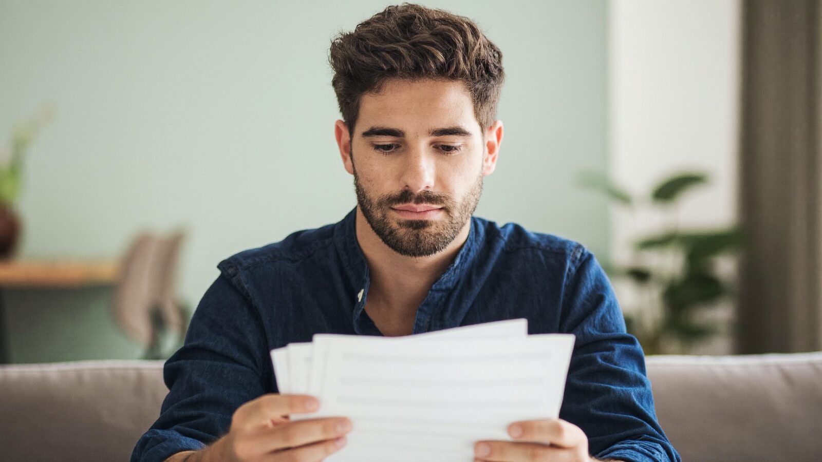 Man in his 30s with thick hair on a living room sofa reviewing paperwork with a calm expression