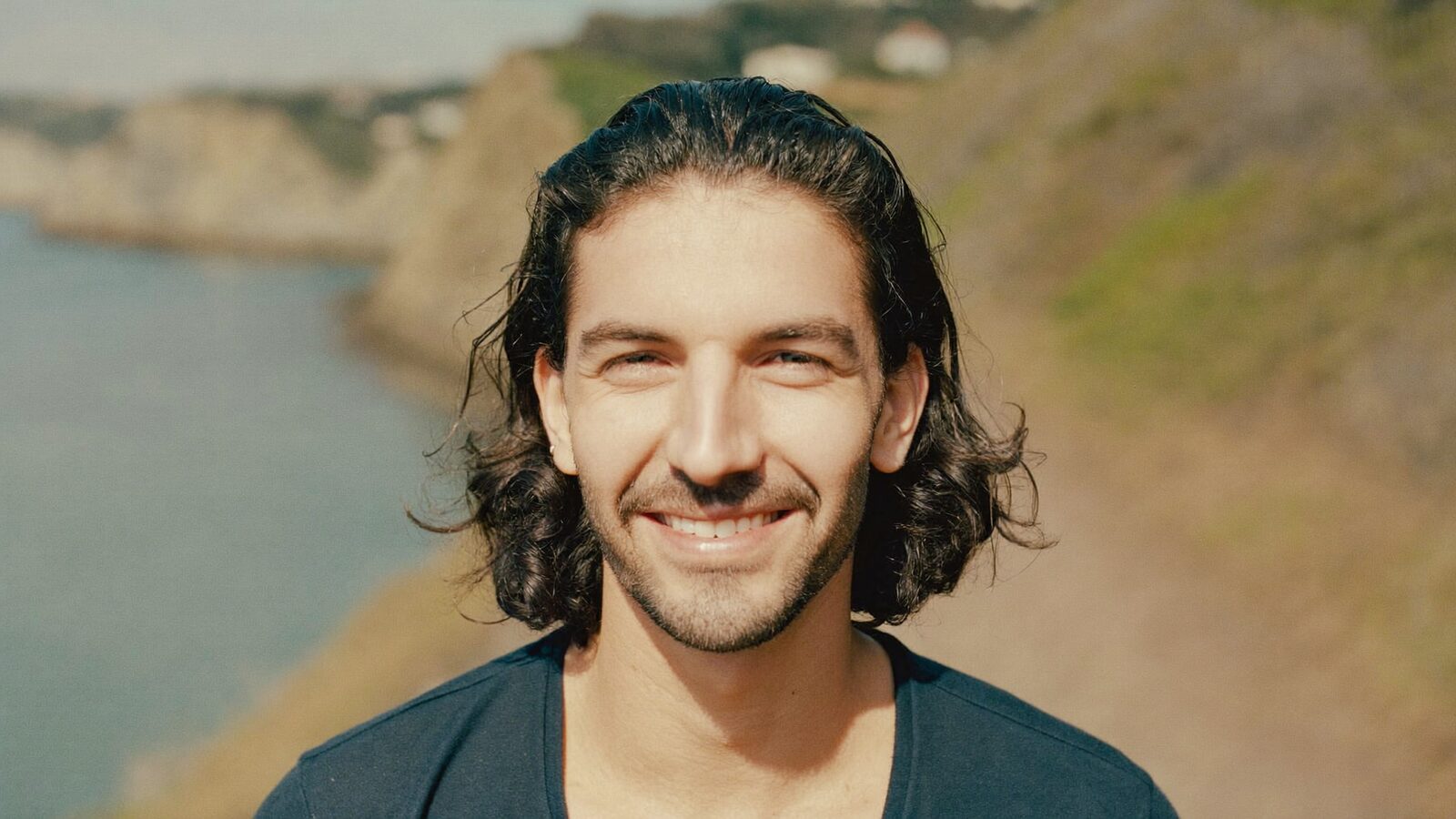 Man in his 30s with shoulder-length wavy hair smiling on a coastal path
