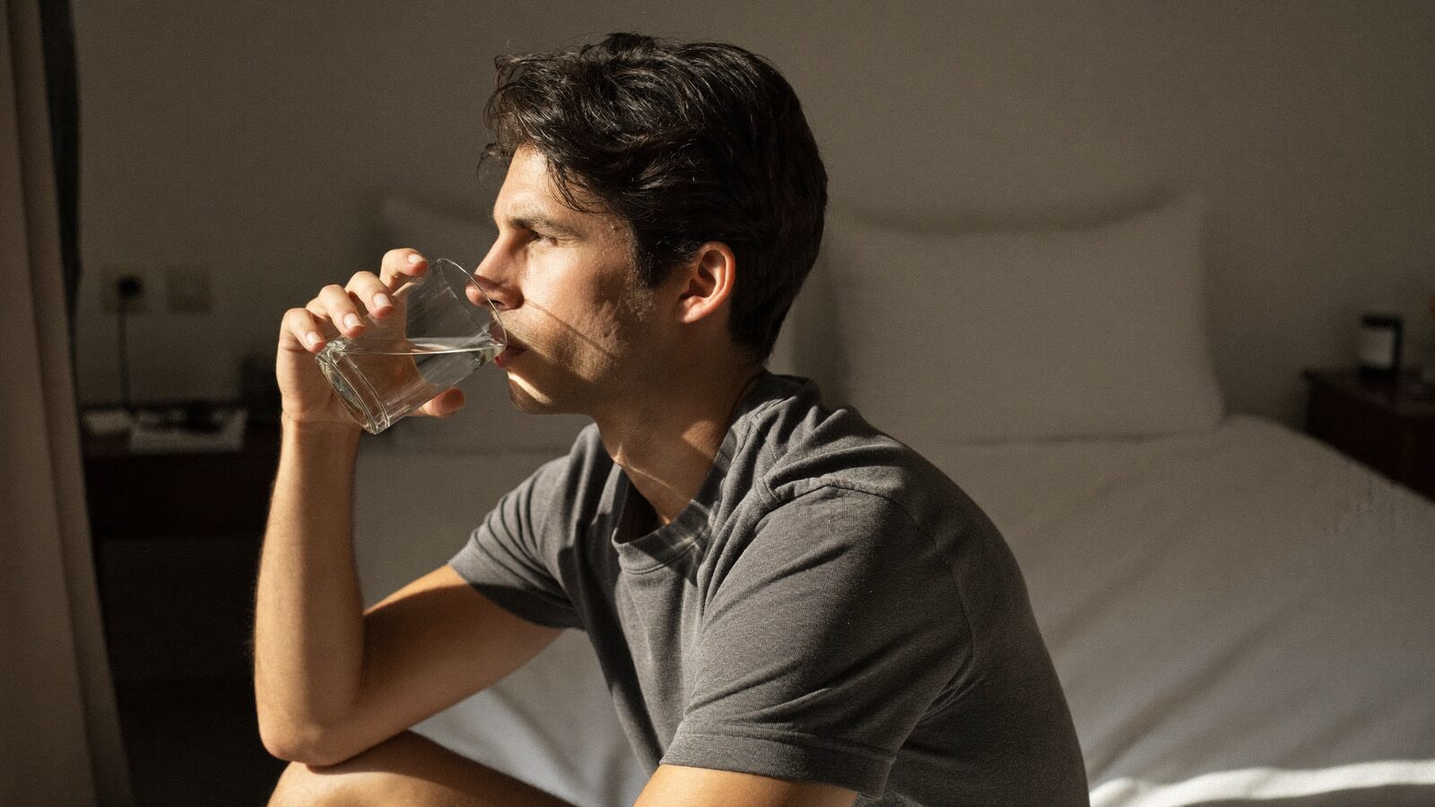 Man in his 30s sitting on his bed in morning sunlight drinking a glass of water