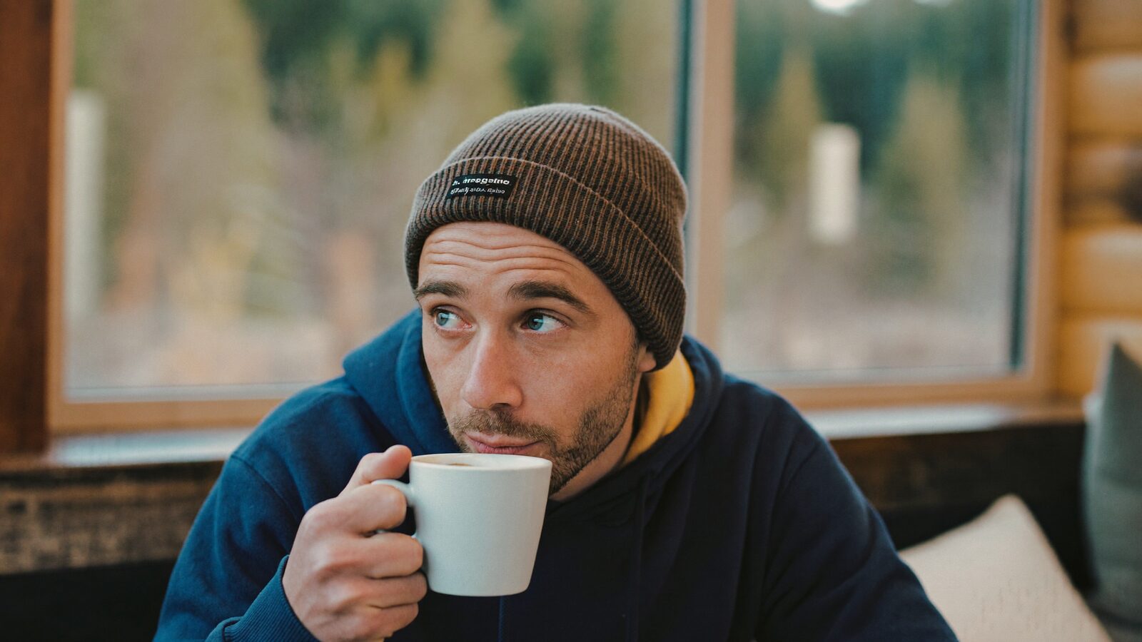 Man in a beanie sipping coffee by a cabin window on a winter morning