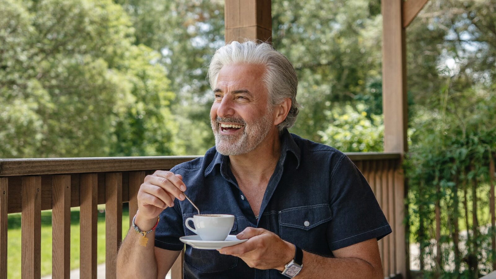 Man in his 60s with thick silver hair laughing on a front porch with a cup of coffee