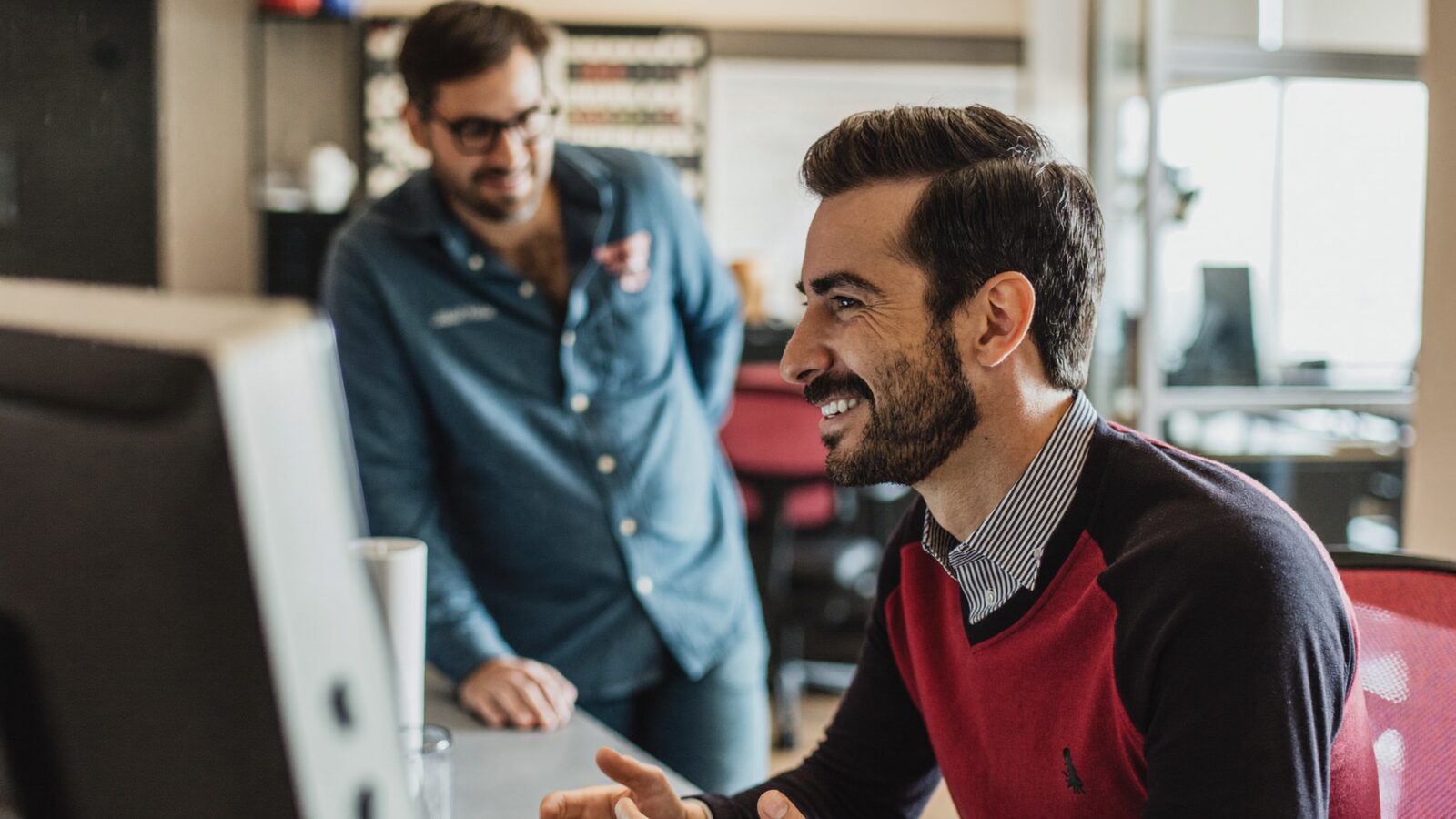 Man in his 30s with neat short hair at a desk in a bright office, talking with a coworker