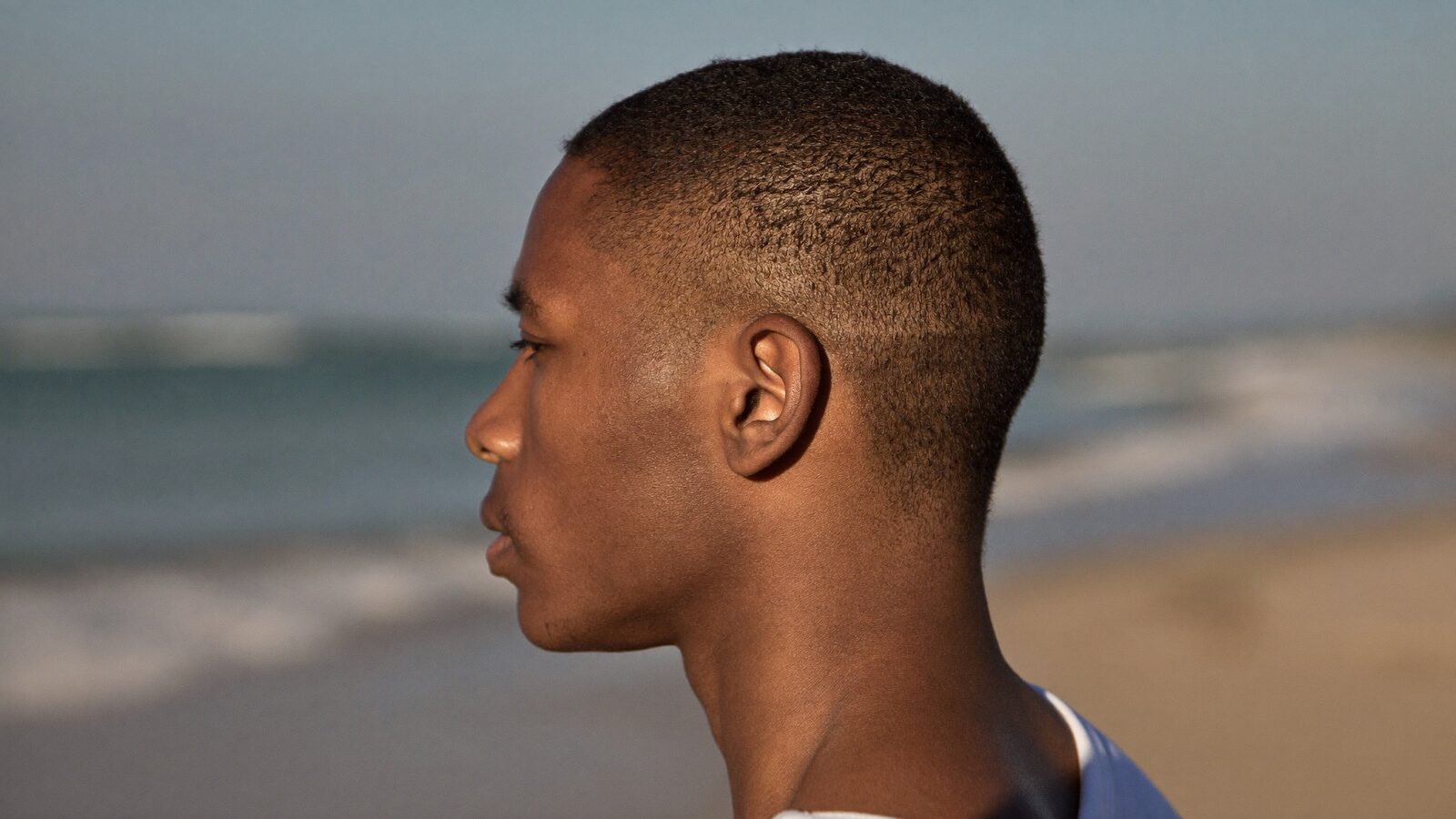 Side profile of a young man with short hair and a clean temple line on a beach at golden hour