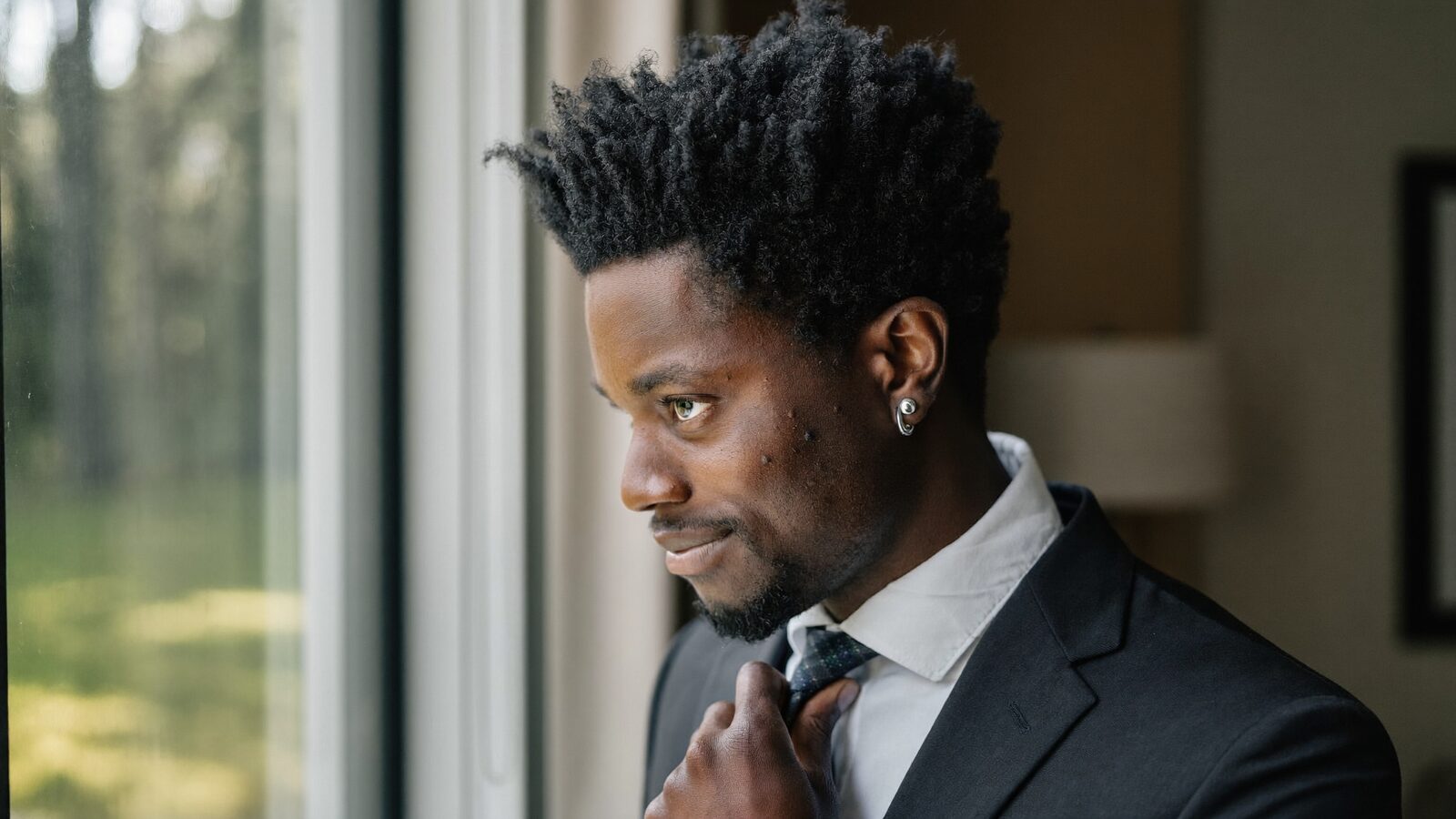 Groom in his 30s adjusting his tie by a window on his wedding morning