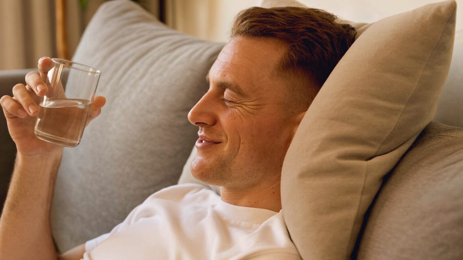 Man resting on a couch against a pillow, smiling and drinking a glass of water