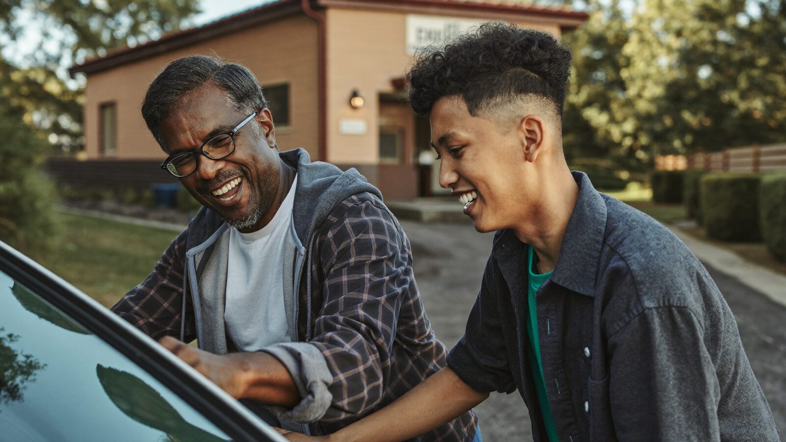 Father in his 50s and son in his 20s laughing together while washing a car in a driveway