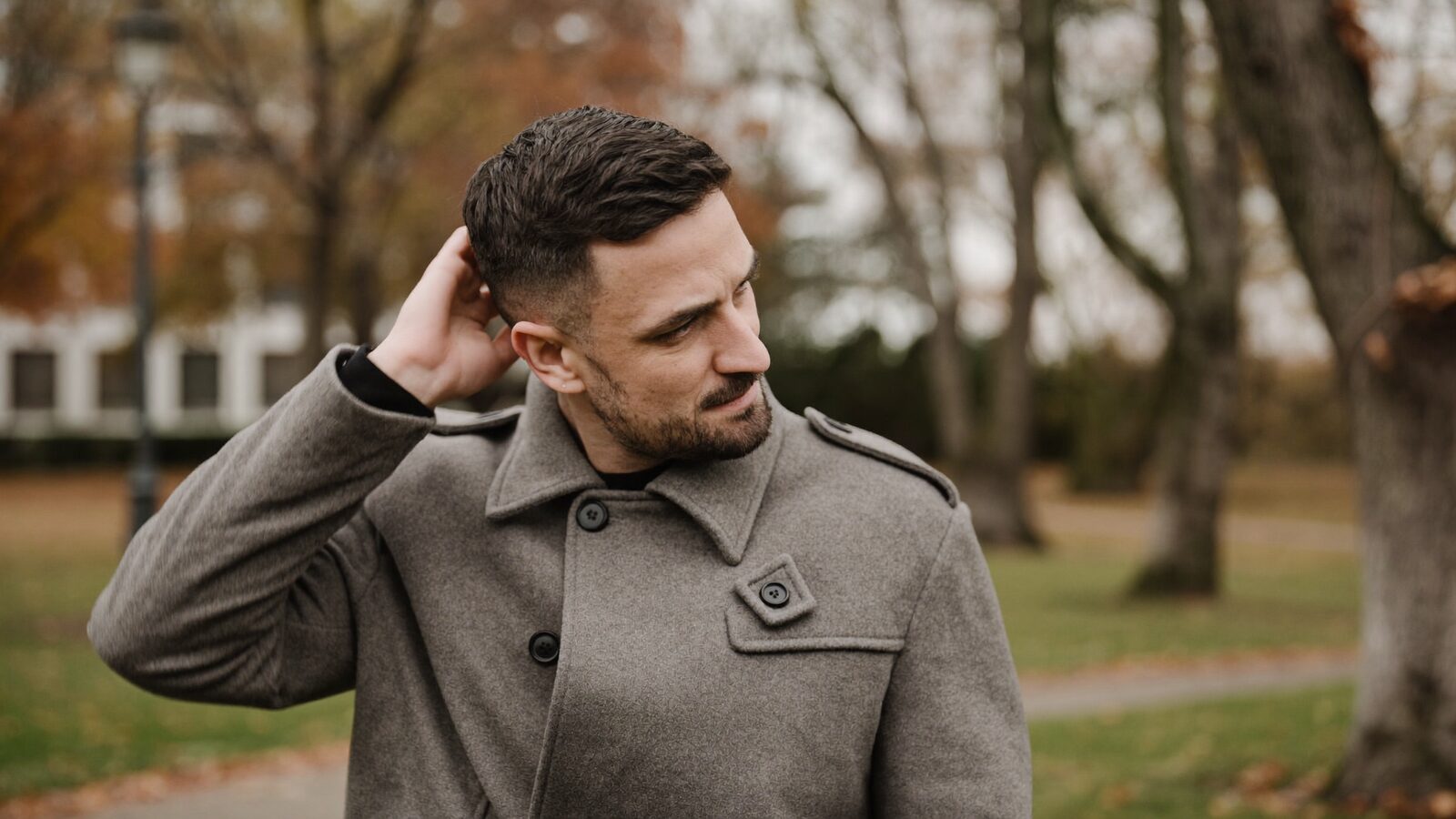 Man in his 30s in a wool coat running a hand through his hair on a windy autumn day