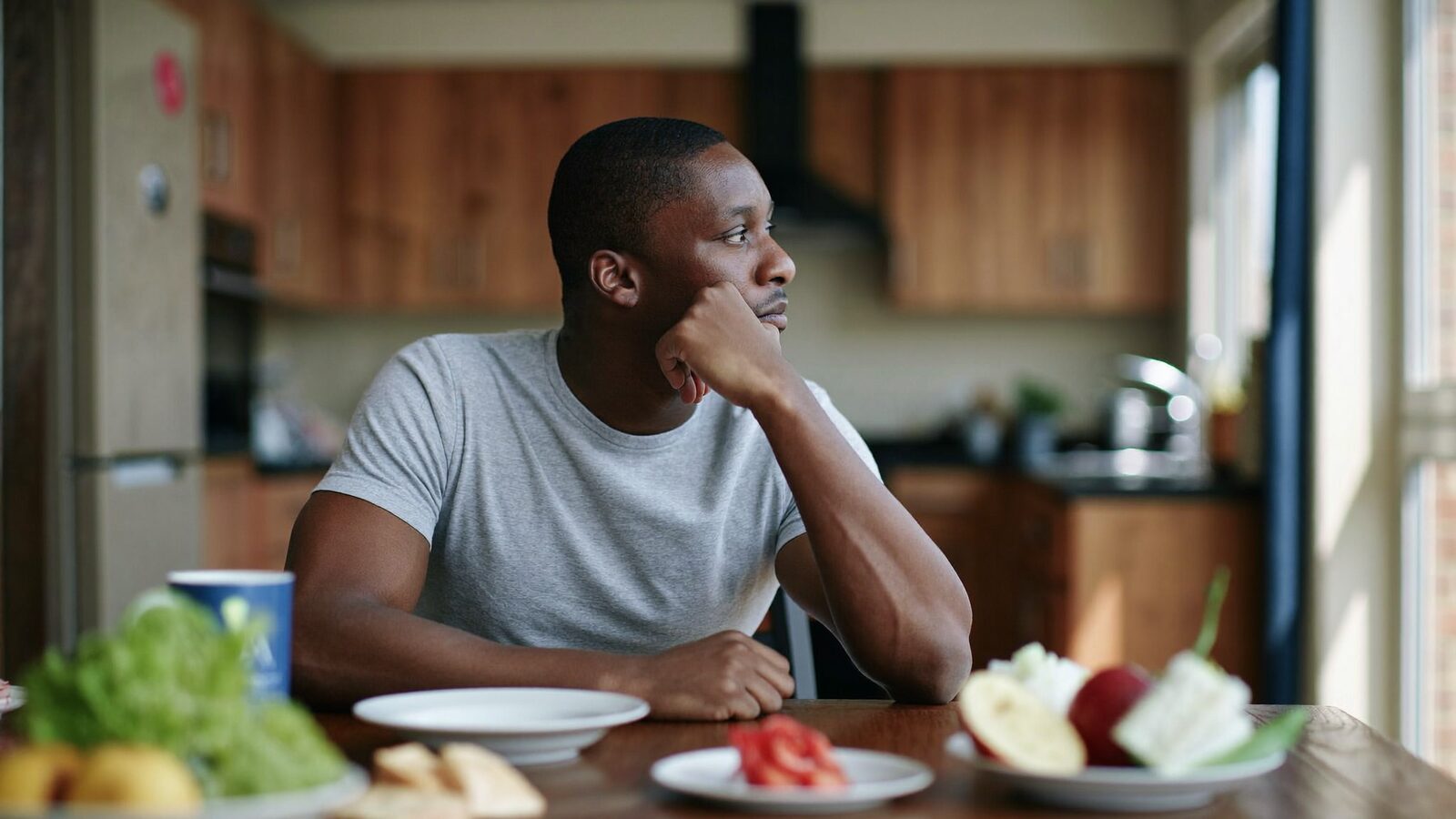 Man in his late 30s with a full natural hairline sitting at a kitchen table at home, looking out the window